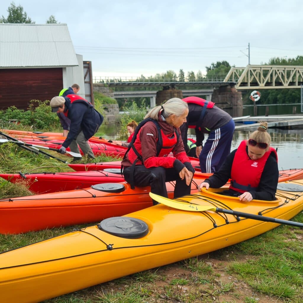 Fyra personer med flytvästar på sig med gula och röda kajaker på strandkanten vid älven.