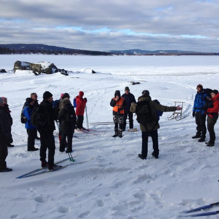 Ett femtontal personer med skidor och sparkar står i snön vid kanten på en istäckt sjö.