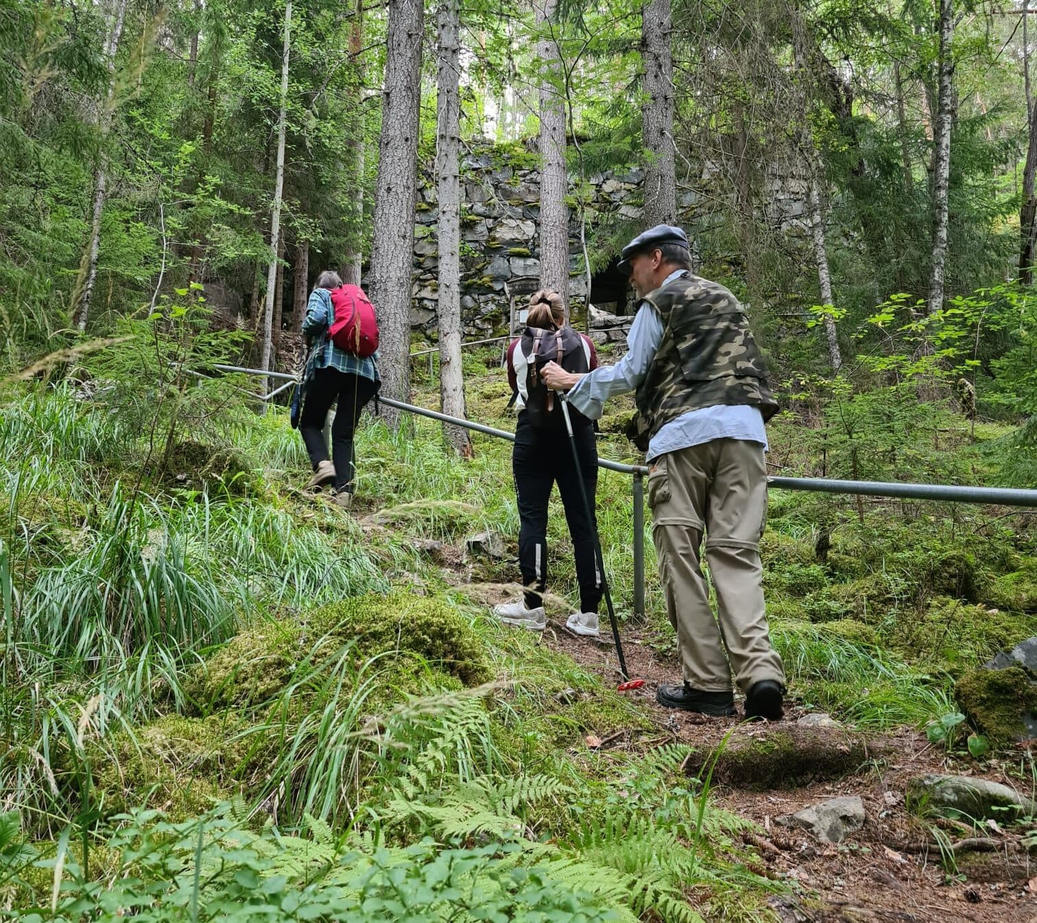Tre personer går uppför en brant stig i vacker gammelskog.