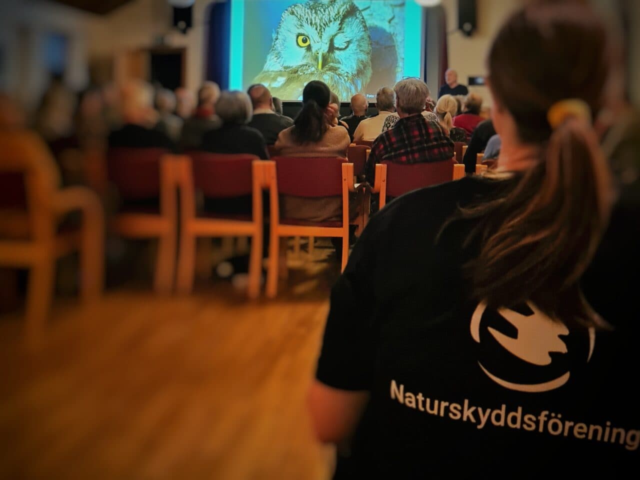 I förgrunden en kvinna med en t-shirt som det står Naturskyddsföreningen på, publik och föreläsare.